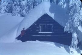 "Colour photo of person by snow-covered cabin"